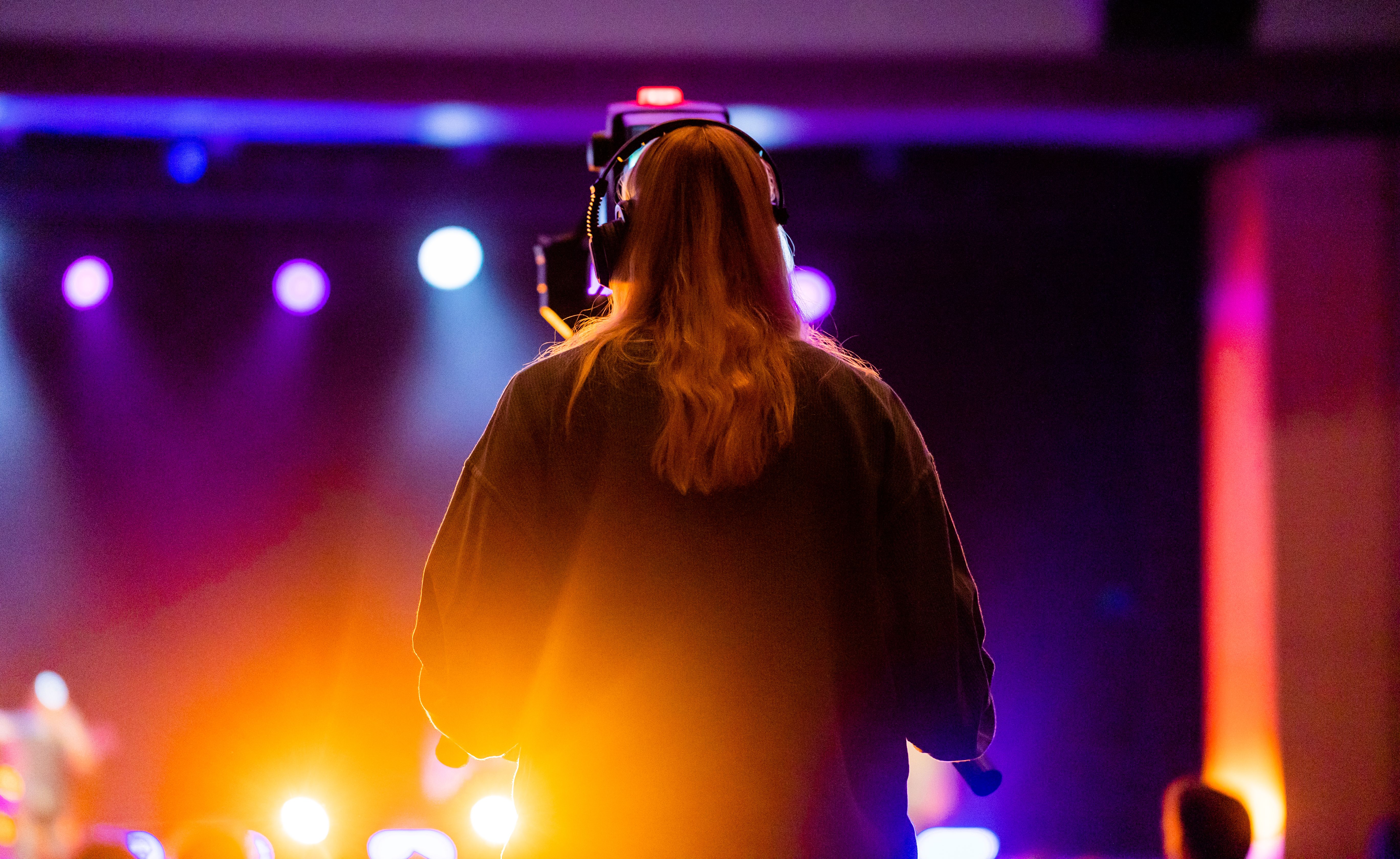 Woman wearing headphones at a concert with vibrant stage lights and colors.