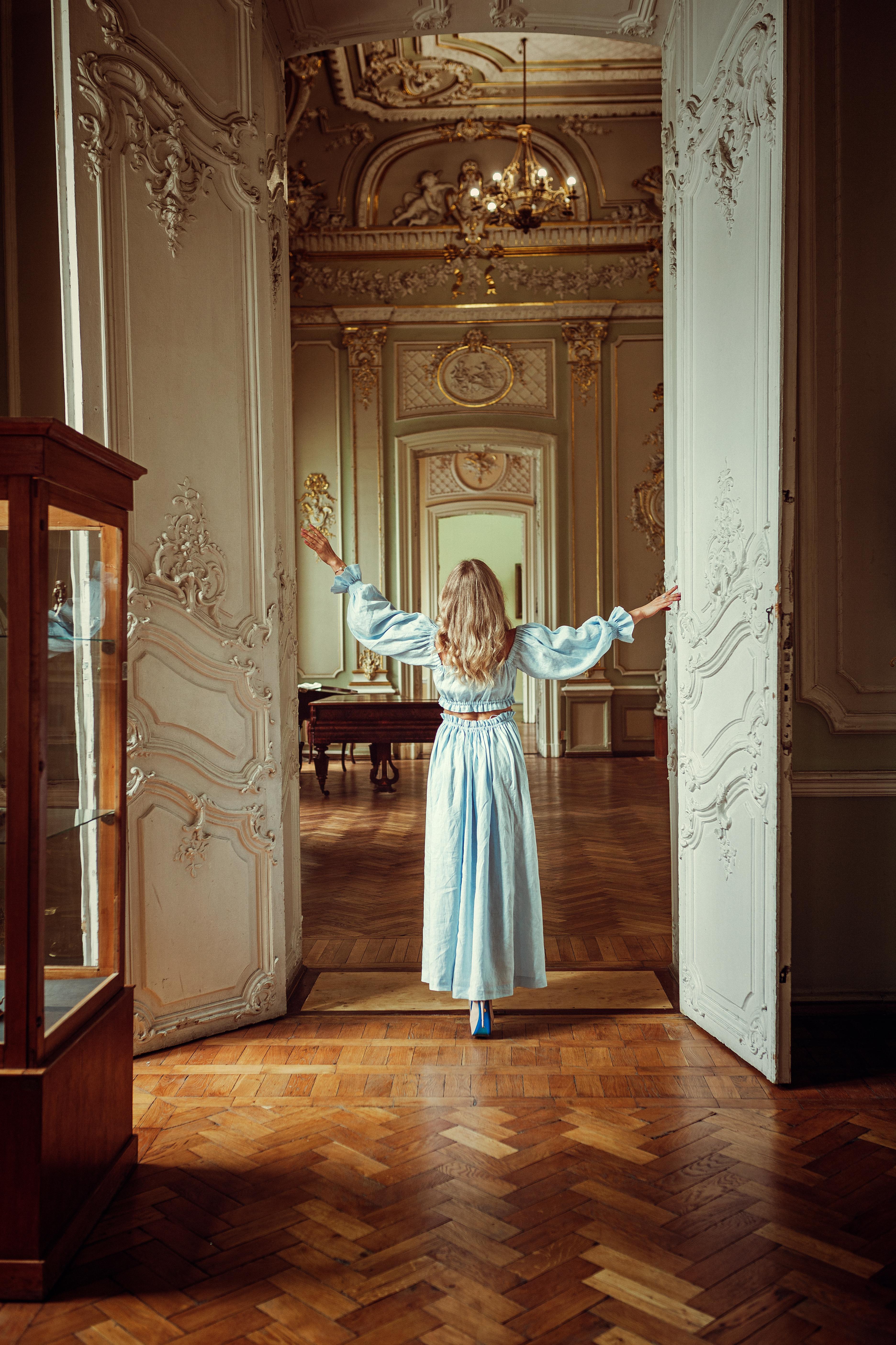 A woman in a light blue dress stands in an ornately decorated room, arms outstretched.