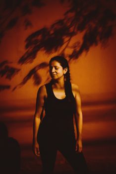 A woman poses outdoors in Brazil, backlit against a warm wall with artistic shadows.
