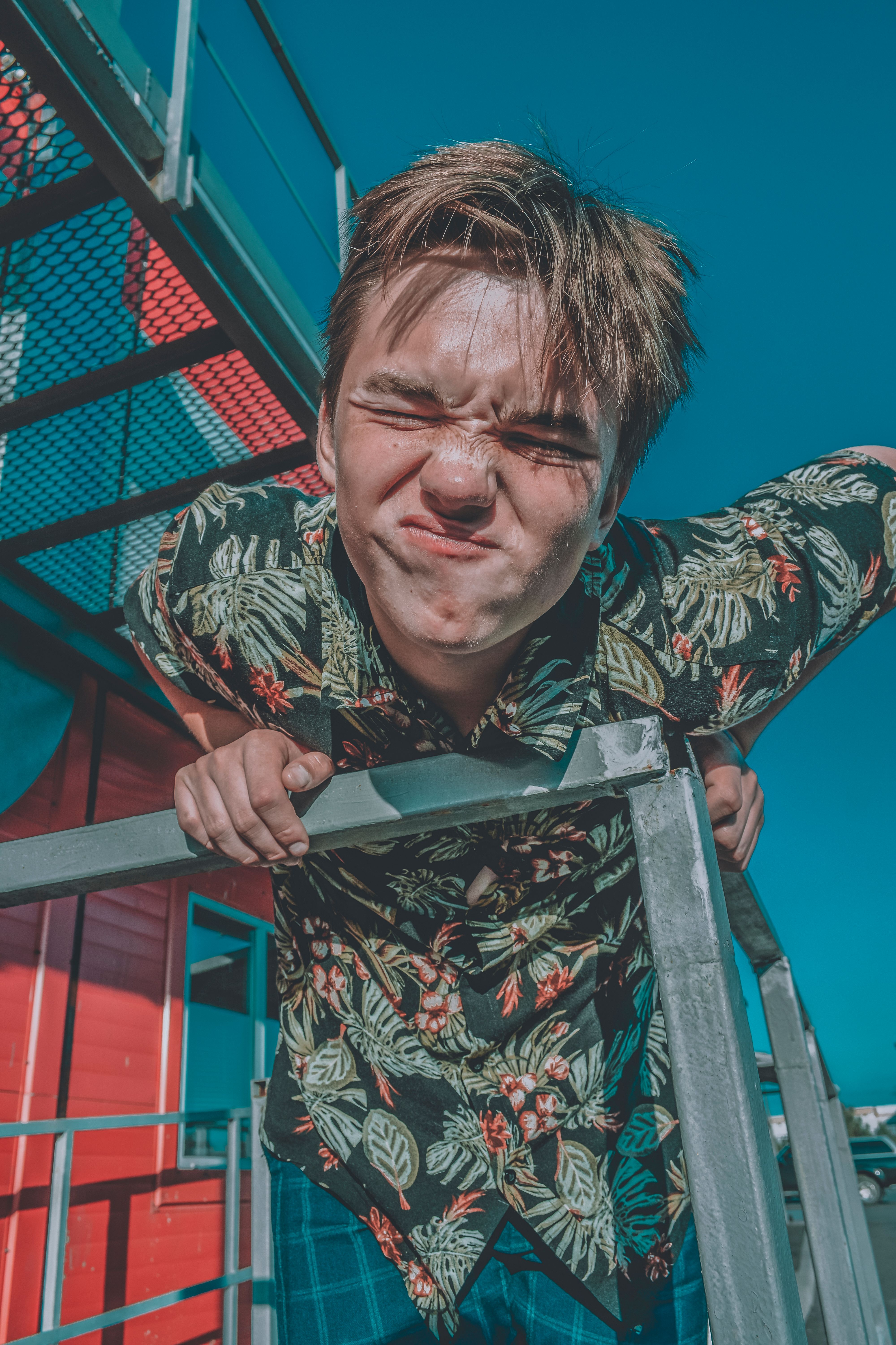 A young man in a floral shirt making a funny face while leaning on a metal railing outside.