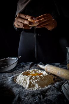 Close-up of hands cracking an egg into flour on a dark surface during baking preparation.