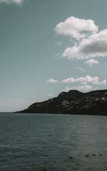 Tranquil coastal scene with rocky hills and soft clouds under a clear sky.