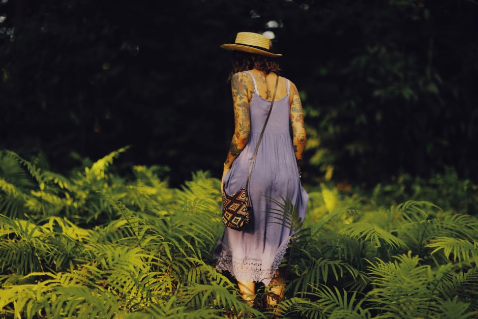 A woman in a hat and summer dress walks through lush green ferns, embracing nature.