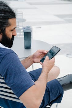 Bearded man in casual attire using smartphone outdoors with coffee tumbler on table.