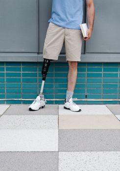 Man with a prosthetic leg holding a book standing against a teal tiled wall.
