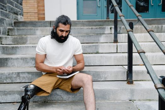 A man with a prosthetic leg is seated on outdoor steps, using a tablet.