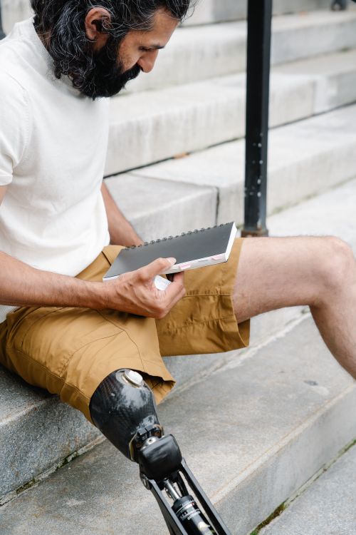 Free Man with a prosthetic leg seated outside, reading a book on steps. Stock Photo