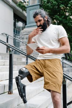 A focused bearded man with a prosthetic leg standing on steps using a tablet.