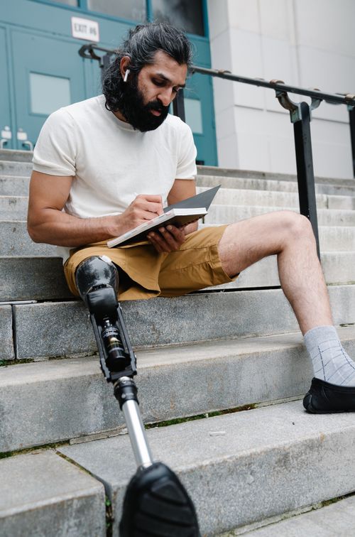 Free Man with a prosthetic leg writing in a journal on steps outside, focused and serene. Stock Photo