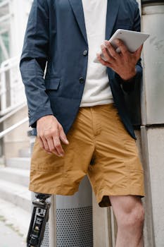 Man in business attire sports shorts and prosthetic leg, holding a tablet outdoors.