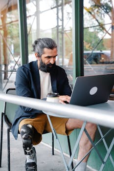 Bearded man with prosthetic leg typing on laptop at outdoor café table.