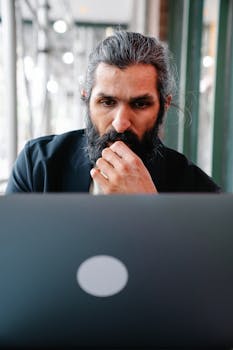 Bearded man deeply focused and working on a laptop in a modern indoor setting.
