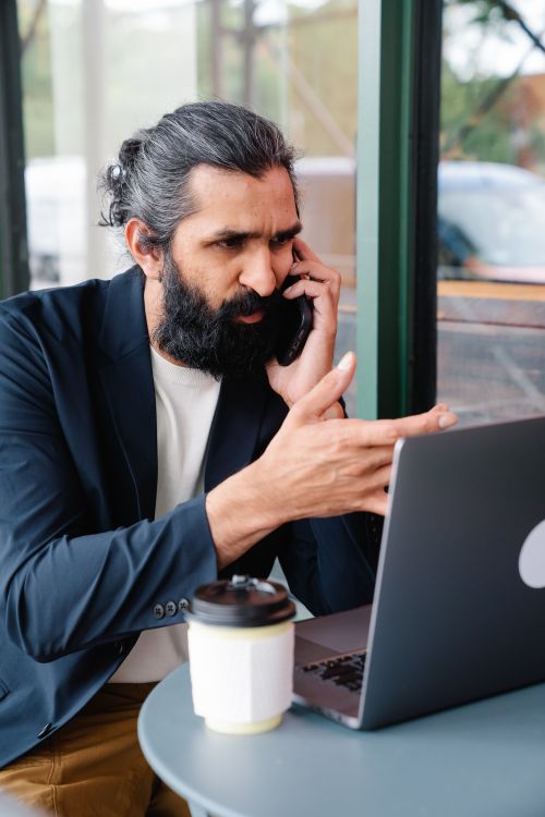 Free Bearded businessman multitasking with phone call and laptop in modern cafe setting. Stock Photo