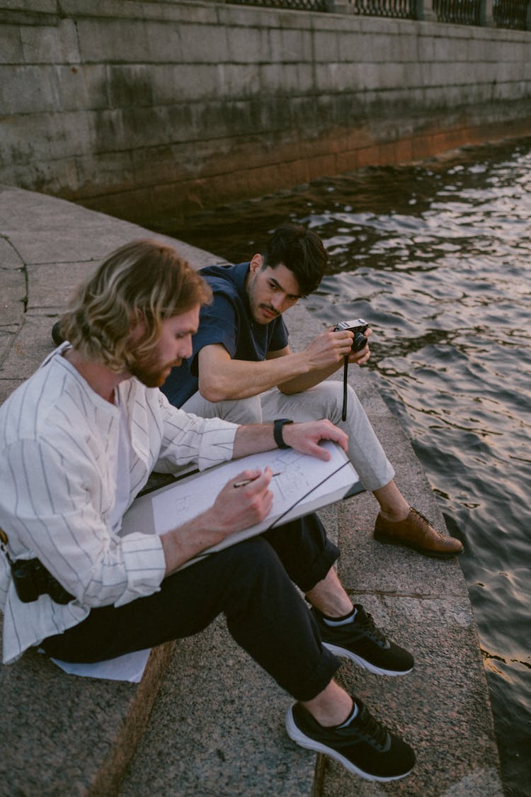 A Man Looking At The Man Sitting Beside Him While Sketching On The Pad