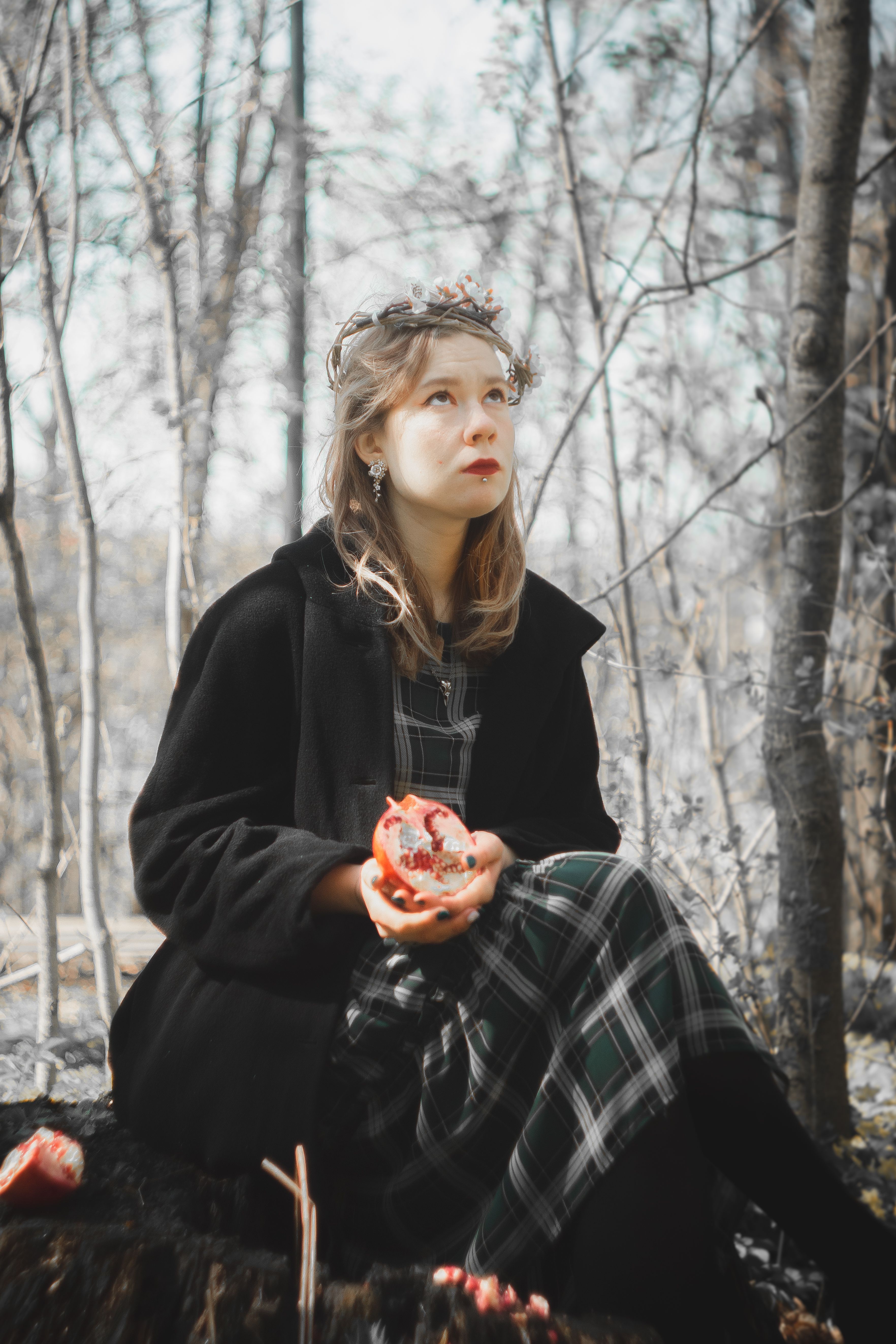 A woman in winter attire sits outdoors holding a pomegranate in a forest setting.