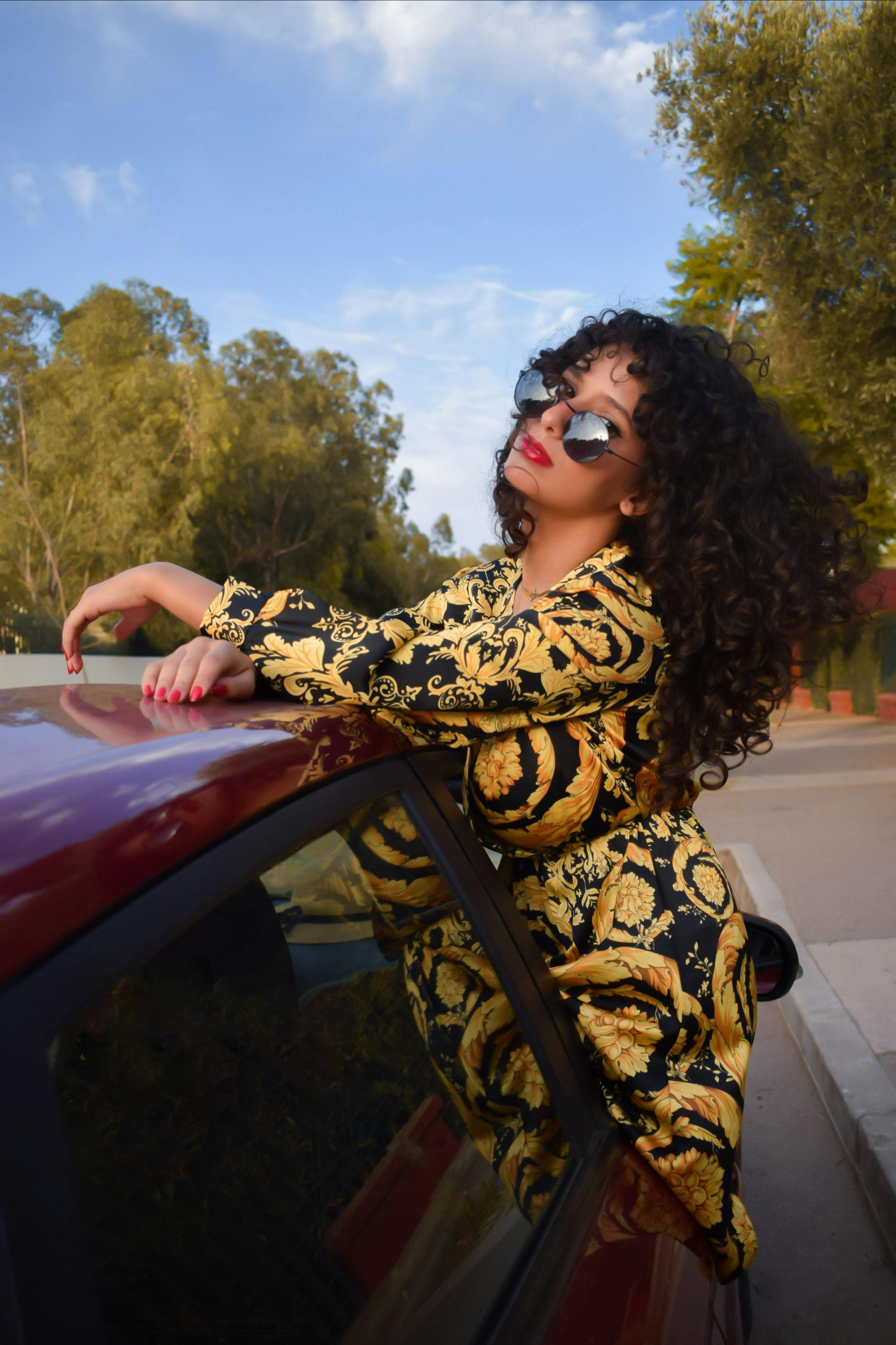 Stylish woman in patterned dress and sunglasses posing on a car in a sunny outdoor setting.