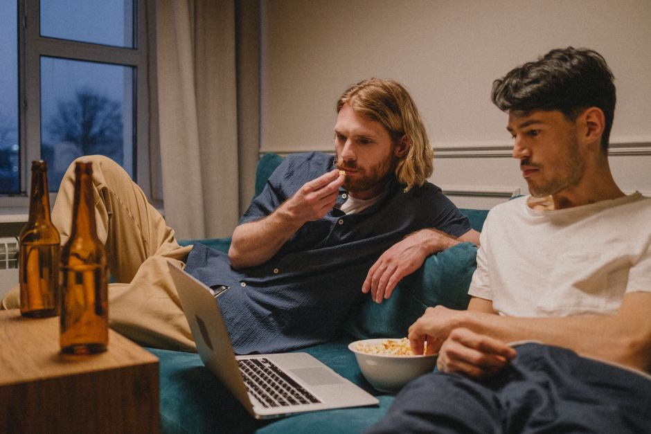 Two friends relaxing at home watching a laptop, enjoying snacks and drinks.