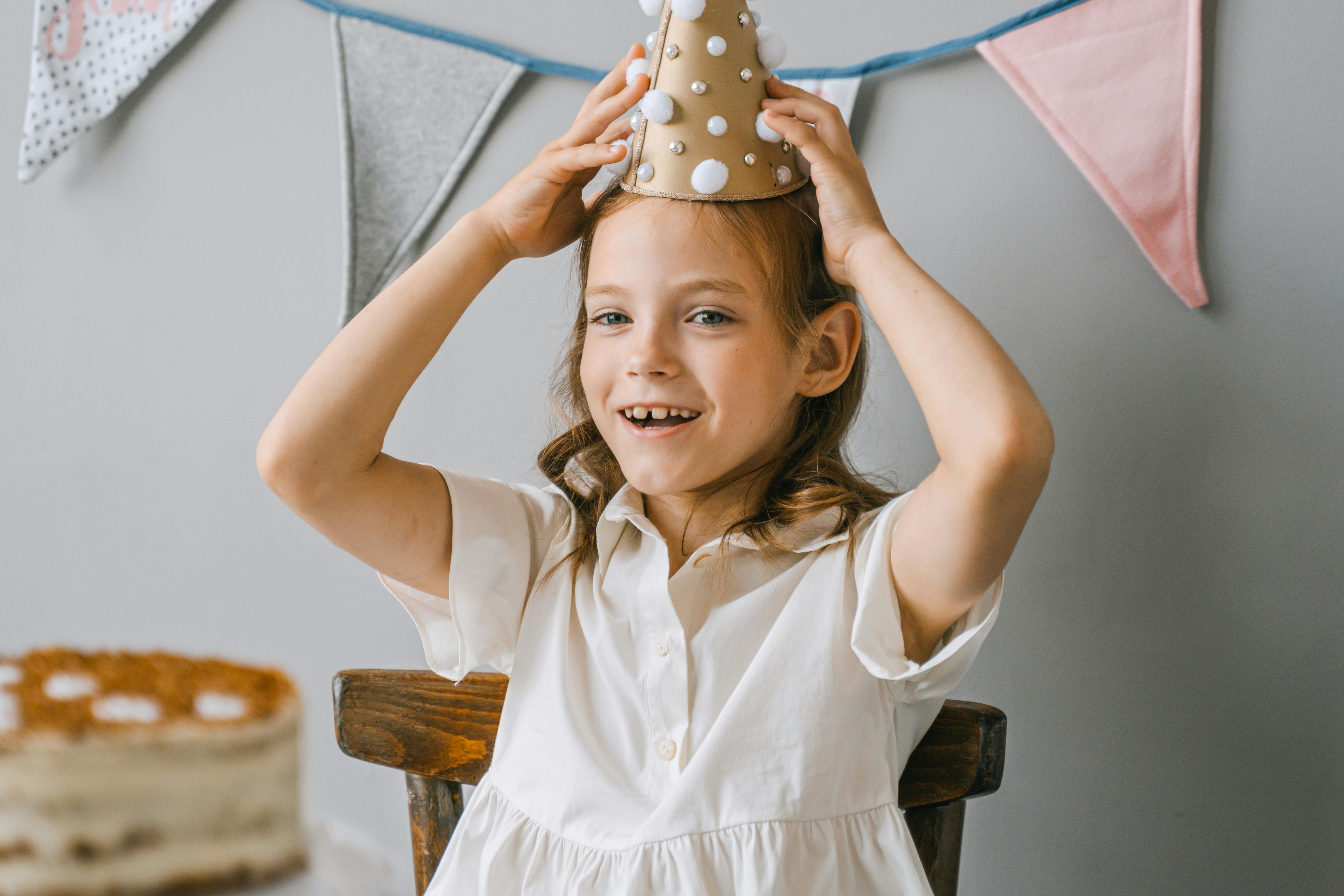 A joyful child at a birthday party wearing a party hat with cake and banners in the background.