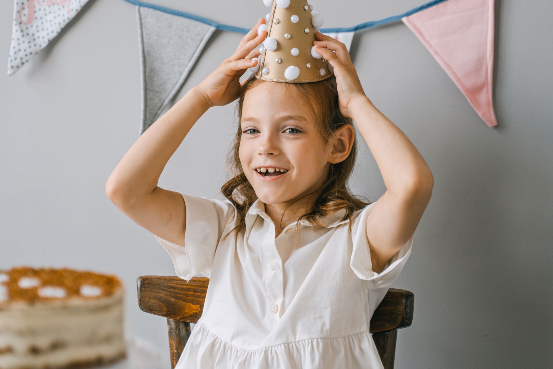 A joyful child at a birthday party wearing a party hat with cake and banners in the background.