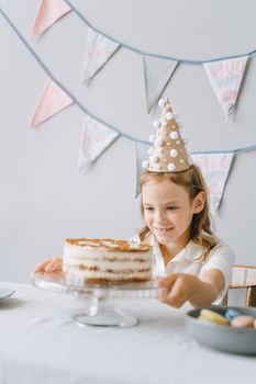 A happy child in a party hat celebrates a birthday with a cake indoors.