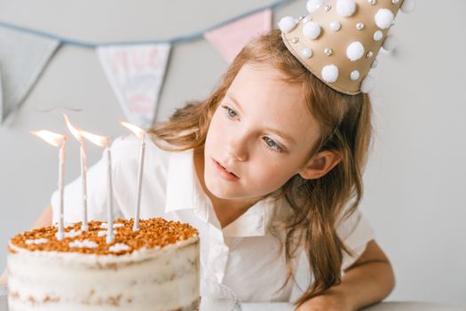 A young girl in a party hat looks at a birthday cake with lit candles indoors.