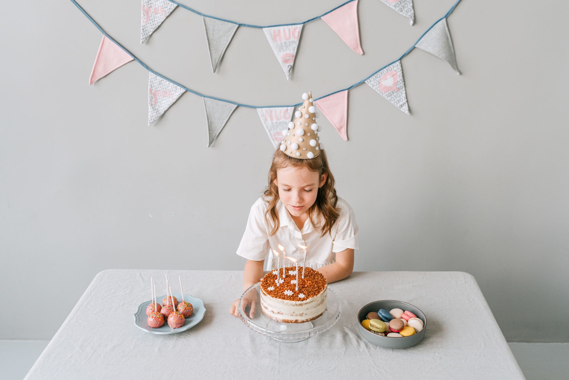 A girl in a party hat making a wish on her birthday cake with macarons on the side.