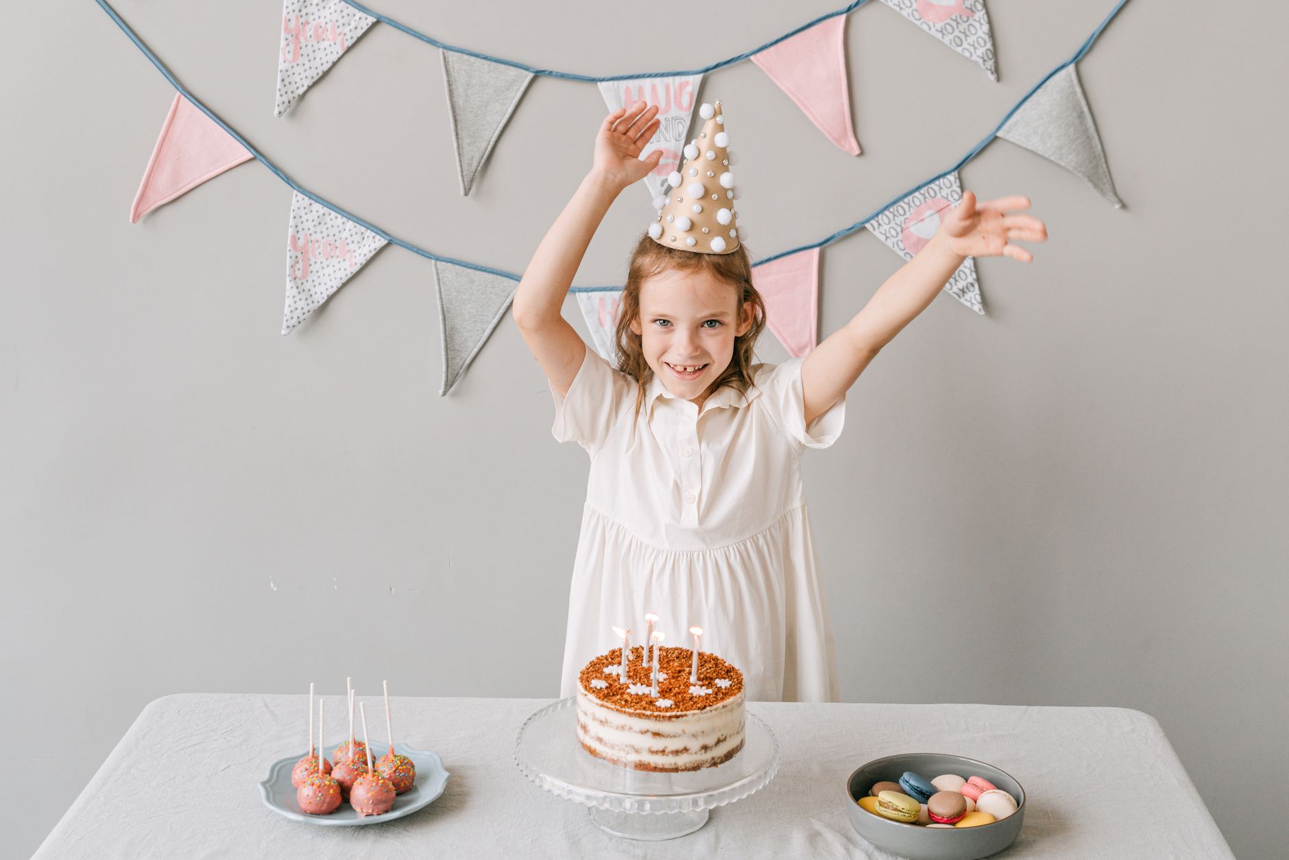 A young girl joyfully celebrates her birthday with a cake, wearing a party hat indoors.