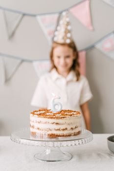 A blurred child in a party hat stands behind a six-year-old birthday cake with a candle.