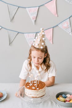 Young girl in party hat about to blow out birthday cake candles indoors.