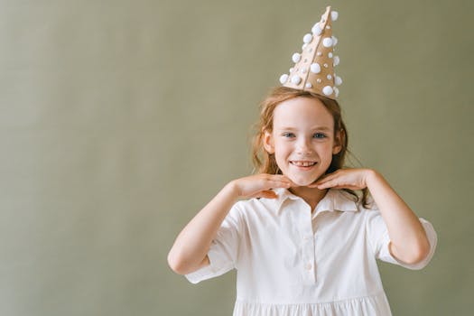 Young girl smiling with a party hat, posing in a white dress against a neutral background.