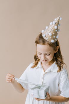A smiling girl with a party hat opens a gift, capturing a joyful birthday moment.