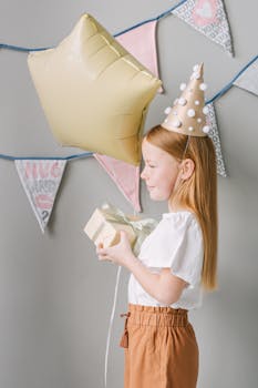 A young girl in party hat holding a gift and balloon at a birthday celebration indoors.