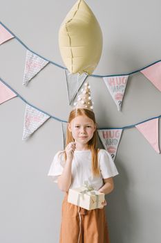 A young girl with red hair holds a gift under party decorations with a balloon and hat.