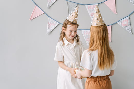 Two girls in party hats exchange gifts at a vibrant indoor birthday celebration.