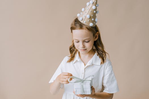 A young girl in a party hat opens a white gift box, wearing a white dress, against a neutral background.
