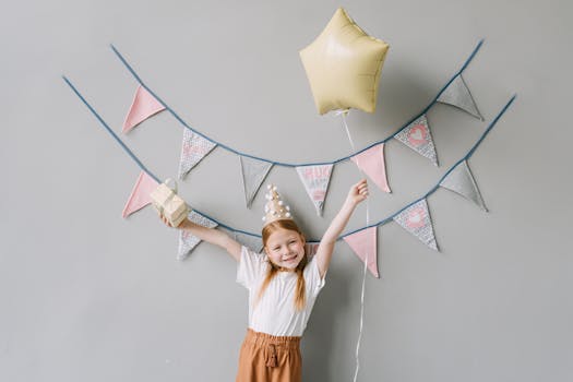 A happy girl in a party hat holding a gift and balloon at a birthday celebration.