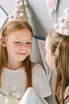 Two young girls in party hats celebrating a birthday indoors with smiles and gifts.