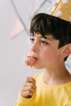 Close-up of a child wearing a party hat and enjoying a colorful lollipop indoors.