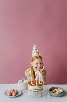 Smiling girl in a gold dress at a birthday party with a cake, lollipops, and macaroons on a pink background.