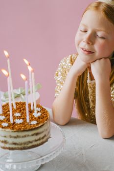 Girl posing with eyes closed beside a birthday cake with lit candles, making a wish.