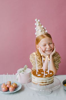 Joyful girl celebrating her birthday, smiling with a cake and party hat on a pink background.