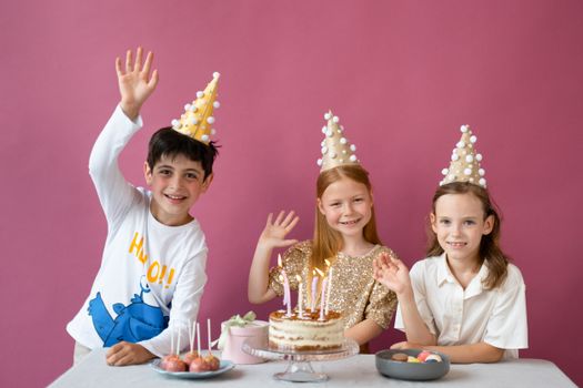 Three happy kids in party hats celebrating a birthday with cake indoors.