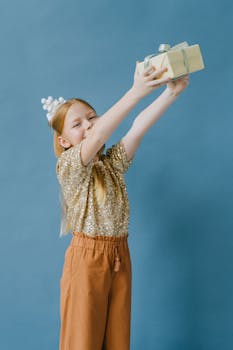Young girl with party hat, holding a gift on a blue backdrop, celebrating joyfully.