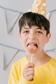 A cheerful boy in a yellow shirt bites a sprinkle-covered cake pop wearing a party hat.