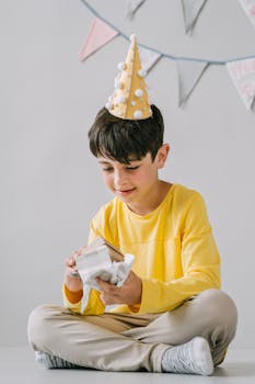 A boy in a party hat joyfully opens a gift during a festive birthday celebration indoors.
