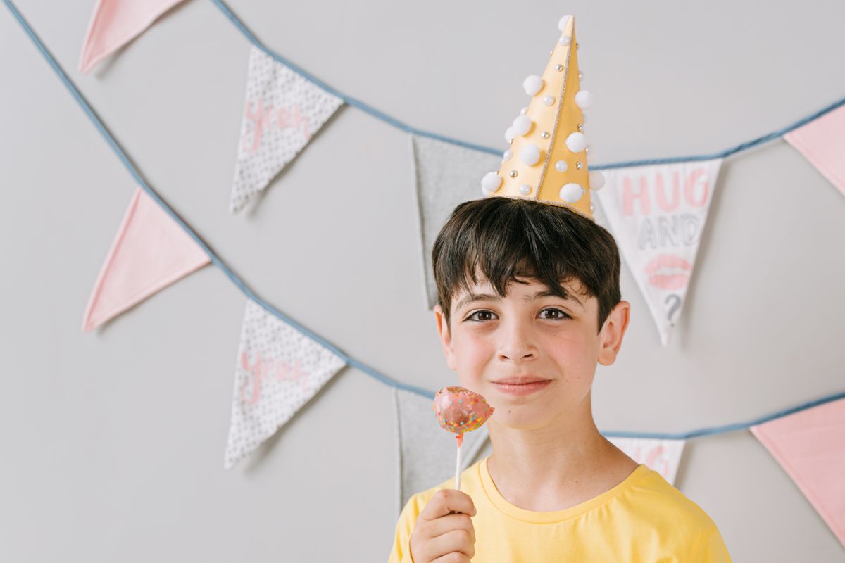 Young boy in a party hat holding a lollipop with festive bunting backdrop.