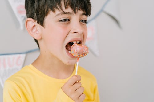 A Close Up Photography of a Boy in Yellow Long Sleeves Eating a Lollipop Cake