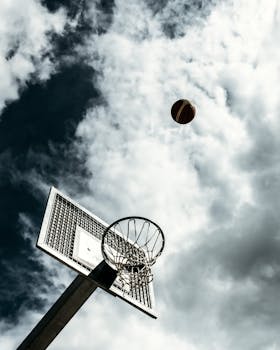 Basketball hoop and ball in midair with cloudy sky background, captured from a low angle.