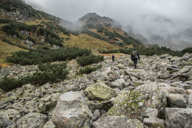 Two hikers navigate a rocky trail in a fog-covered mountainous landscape.