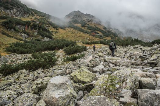Two hikers navigate a rocky trail in a fog-covered mountainous landscape.